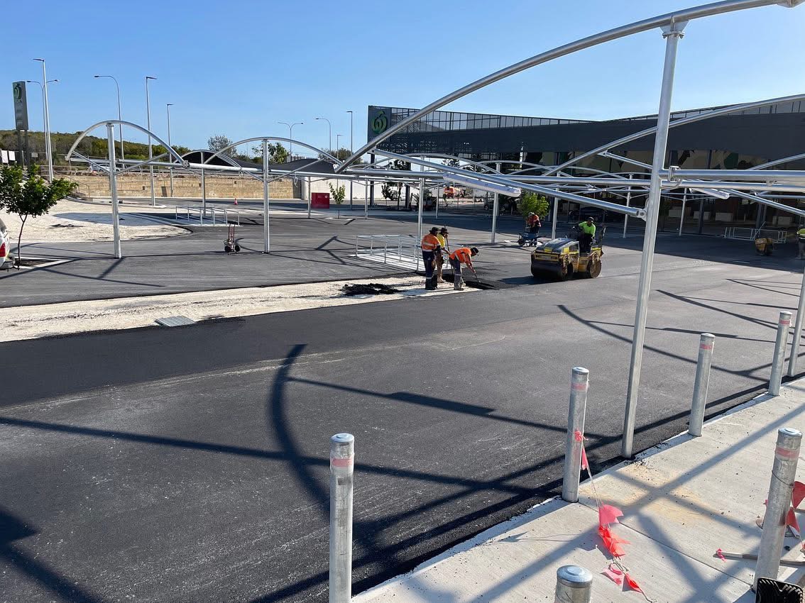 YongaCivil crew laying and compacting asphalt in a Woolworths shopping centre car park with curved shade sail canopies